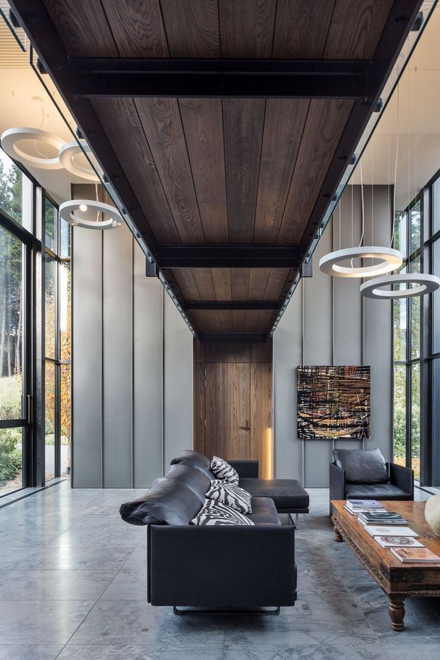 A dramatic contemporary living room with double-height proportions and floor-to-ceiling black-framed glazing on both sides. A dark timber-clad overhead ceiling element with exposed black steel framing leads toward a tall timber door and creates a strong axial composition. The room features black leather sofas and armchairs, sculptural circular pendant lights, a rustic carved timber coffee table, large-format grey stone flooring, and abstract wall art. The space is refined, moody, and highly architectural, with abundant natural light and views of greenery outside.