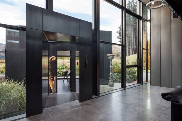 A dramatic contemporary entry hall with floor-to-ceiling black-framed glazing, dark vertical wall panels, and large-format grey tiled flooring. A sculptural doorway and artwork lead into an adjoining interior space, while landscaped grounds and distant hills are visible through the glazing. The space is bright yet moody, with strong geometric lines and a refined, minimalist character.