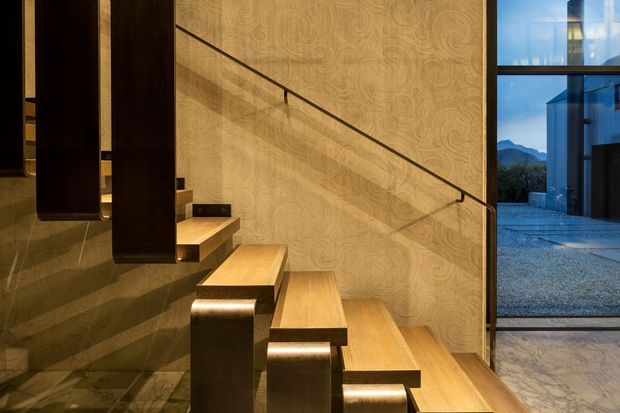 A contemporary interior stairway with floating timber treads supported by dark rectangular structural elements. The staircase runs alongside a patterned feature wall with a slim dark handrail, while large glazed doors or windows reveal an outdoor courtyard and distant mountain landscape. Stone or marble-look flooring and warm integrated lighting add a refined, dramatic atmosphere.