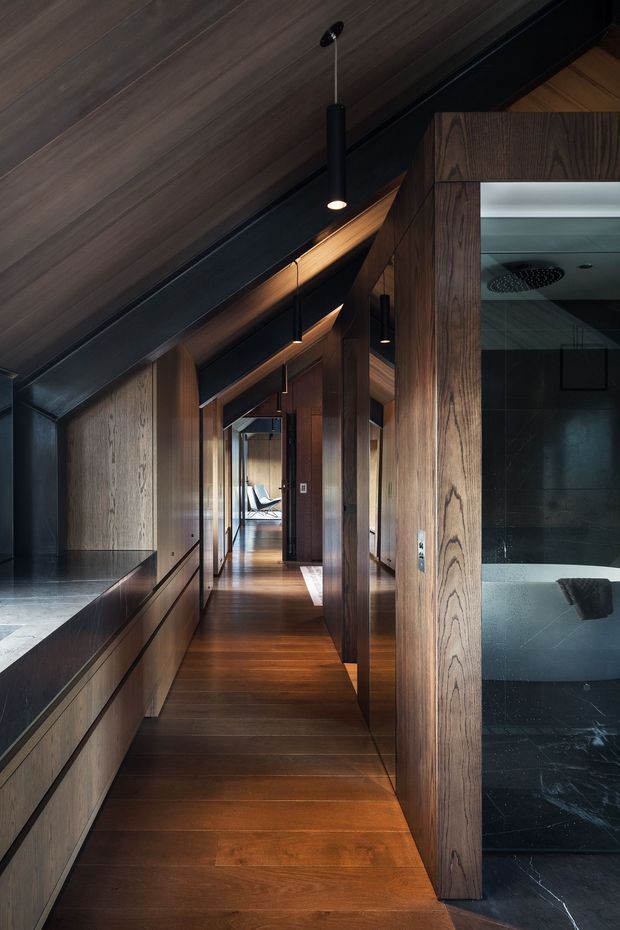A narrow, sophisticated residential hallway lined with dark timber wall panelling and matching timber flooring. The corridor has a dramatic sloped timber ceiling with exposed black structural beams, recessed and slim black pendant lighting, and concealed built-in cabinetry along the left side. A large timber-framed opening on the right reveals a dark tiled bathroom with a freestanding bathtub and ceiling shower fixture. Glazing and doorways lead toward a brighter room at the end of the hall.