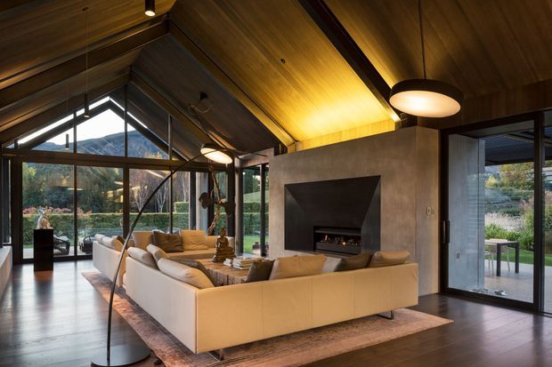 A warm, sophisticated living room with a dramatic pitched timber ceiling and extensive floor-to-ceiling glazing. A large cream sectional sofa is arranged around a freestanding concrete fireplace wall with a recessed dark firebox. The room opens to a landscaped garden through glazed doors, with dark flooring, layered rugs, sculptural pendant lighting, and a visible outdoor terrace beyond.