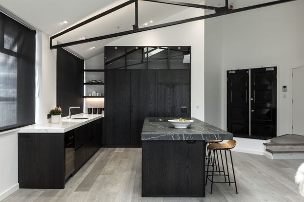 Contemporary open-plan kitchen with dark timber cabinetry, a large veined stone island, integrated shelving, black-framed internal glazing, and a black feature entry door. Light timber-look flooring and white walls create a bright, high-contrast interior.