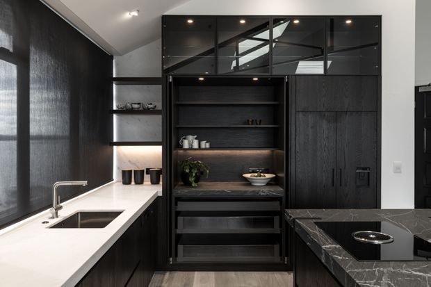 A sophisticated dark-toned kitchen with black timber cabinetry, integrated open shelving, a white stone island or benchtop, and a contrasting dark marble island with a built-in cooktop. Floor-to-ceiling cabinetry includes glass-fronted upper storage, while a large skylight or roof window brings natural light into the space. The design has a refined, high-contrast contemporary feel.