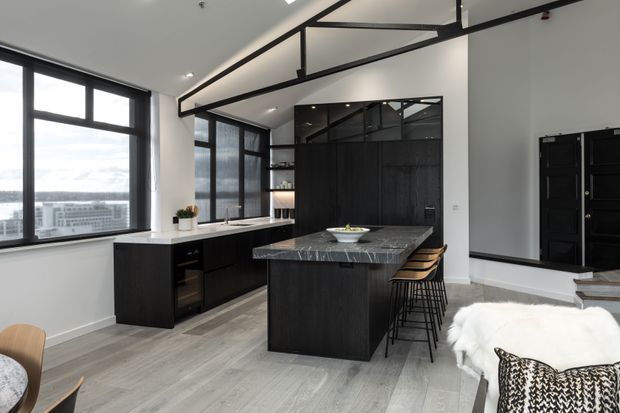 A contemporary open-plan kitchen and living area in a high-rise setting. The kitchen features dark wood-toned cabinetry, a large grey stone island with bar seating, black-framed floor-to-ceiling windows, and a black-framed internal glass partition. A small portion of the living and dining areas is visible in the foreground.
