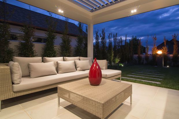 Covered outdoor lounge terrace at dusk with a large woven sectional sofa, matching square coffee table, pale tiled flooring, and a red decorative vase. The space has a white structural frame, illuminated slatted pergola roof, and open views to a landscaped garden with stepping stones and feature lighting.
