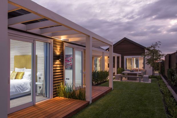 Twilight exterior view of a contemporary single-storey residential development arranged around a narrow landscaped lawn. The image highlights timber-clad facade sections, extensive glazed sliding doors, covered pergola walkways, timber decking, warm integrated lighting, and a small outdoor dining area. Bedrooms and interior furnishings are visible through the glazing.