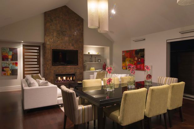 Open-plan living and dining room with a dark timber dining table, upholstered dining chairs, a white sofa, and a stone-clad fireplace wall incorporating a television and firebox. Warm pendant lighting, recessed shelving, timber flooring, and colourful artwork create a refined contemporary interior.