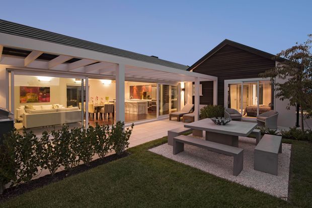 Contemporary single-storey home with a dark gabled pavilion connected to a light, flat-roofed glazed living wing. The image looks across a landscaped courtyard with an outdoor dining table and bench seating, while the open interiors reveal a living room, dining area and kitchen. Strong indoor–outdoor connection through extensive sliding glazing and a covered pergola.