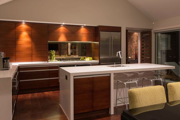 A warm, contemporary kitchen with extensive dark timber cabinetry, integrated stainless-steel appliances, a large white island with a sink and breakfast bar, and clear acrylic bar stools. Under-cabinet and display lighting highlight the timber joinery, while a glazed door and adjacent doorway provide additional depth.