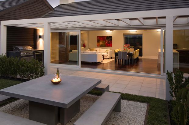 A contemporary home viewed from a landscaped courtyard, with a covered outdoor entertaining area opening into a warm, open-plan living and dining room. The exterior features dark horizontal cladding, a light pergola structure, large sliding glass doors, tiled paving, built-in barbecue, and a concrete dining table with bench seating and a small fire feature.