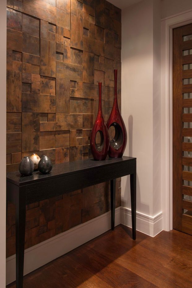 A warmly lit interior hallway or entryway featuring a dramatic patchwork copper-toned metal feature wall, dark console table, sculptural red decorative vases, and rich dark timber flooring. A timber interior door with horizontal inset glass panels is visible on the right, with white painted trim and skirting providing contrast.