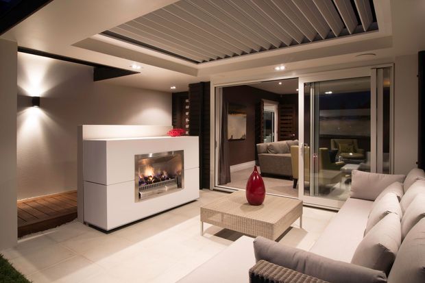 Contemporary covered outdoor lounge or alfresco entertaining area with a white built-in fireplace, pale tiled flooring, a large upholstered sectional sofa, coffee table, and sliding glass doors opening to an interior living room. The ceiling features a prominent louvred or slatted detail, while warm integrated lighting creates an inviting evening atmosphere.