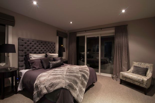 A sophisticated, dark-toned bedroom with a large tufted upholstered headboard, layered charcoal and mauve bedding, and a plush faux-fur throw. Full-height curtains frame glazed doors, while bedside tables, table lamps, recessed spotlights, and a patterned accent chair complete the refined interior.