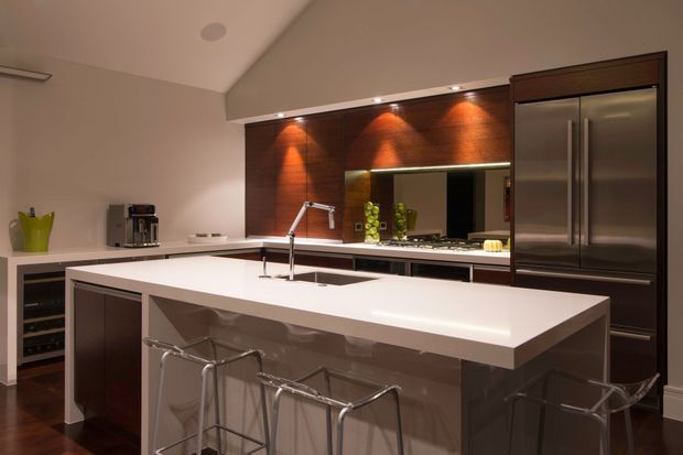 Contemporary kitchen with a large white island, integrated sink and chrome tap, dark timber cabinetry, stainless-steel refrigerator, glossy splashback, and warm recessed accent lighting. A breakfast bar with several chrome stools occupies the foreground.