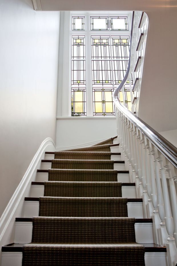 A bright, elegant residential stairway viewed looking upward. The staircase has a dark patterned stair runner, white painted balustrades and moulded trim, dark timber stair edges, and a polished timber handrail. A large multi-panel leadlight-style window with geometric stained-glass detailing illuminates the landing, creating a refined heritage character.