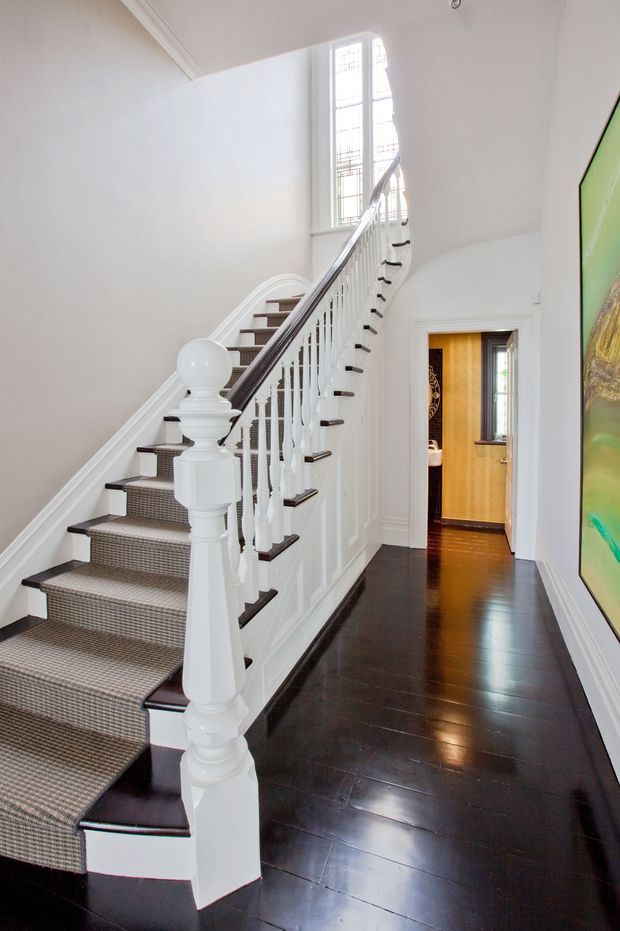 Bright, traditional-style residential stair hall with a carpeted staircase, white painted turned balusters and newel posts, dark polished timber flooring, decorative wall mouldings, and tall leaded-glass windows. A doorway opens to a warm-toned adjacent room, while a large colourful artwork is visible along the right wall.