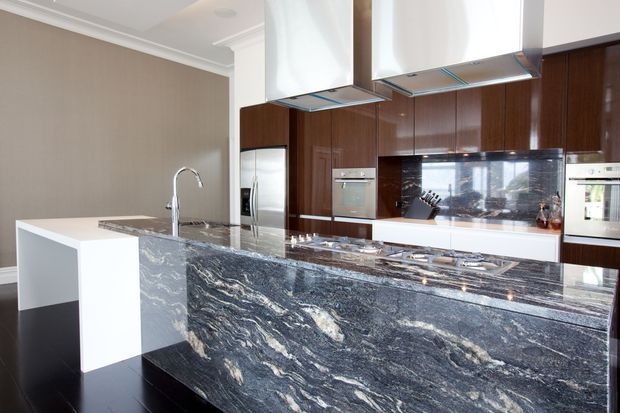 A sophisticated contemporary kitchen featuring a large waterfall island with dramatic dark veined stone, white cabinetry, rich dark timber wall cabinetry, stainless-steel appliances, and dual stainless-steel rangehoods. The space has a warm neutral palette and polished, high-contrast finishes.