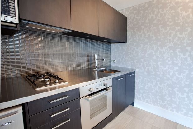 Compact contemporary kitchen with dark timber-look overhead and lower cabinetry, stainless-steel benchtop and integrated gas cooktop, stainless oven, and a metallic linear-tile splashback. The kitchen is set against a pale patterned feature wall and light timber-look flooring.