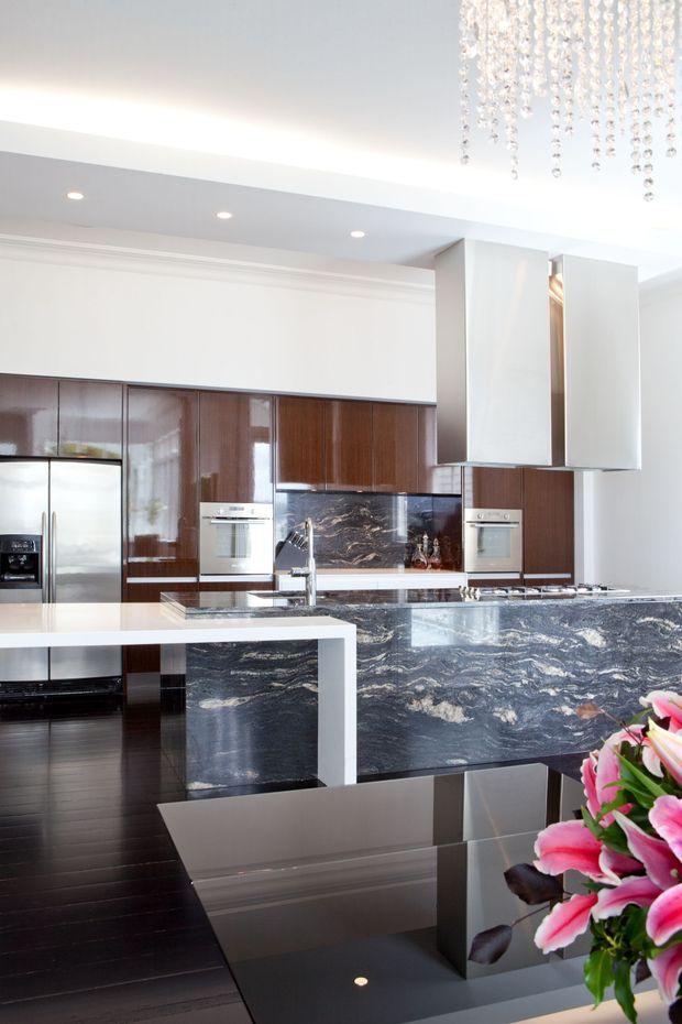 Bright, high-end contemporary kitchen with dark glossy timber cabinetry, stainless-steel appliances, a large sculptural stone island and matching stone splashback. The kitchen includes a prominent white waterfall-edge peninsula, dark timber flooring, recessed ceiling lights, and a crystal pendant visible at the top right. A reflective dark dining or breakfast table and flowers occupy the foreground.