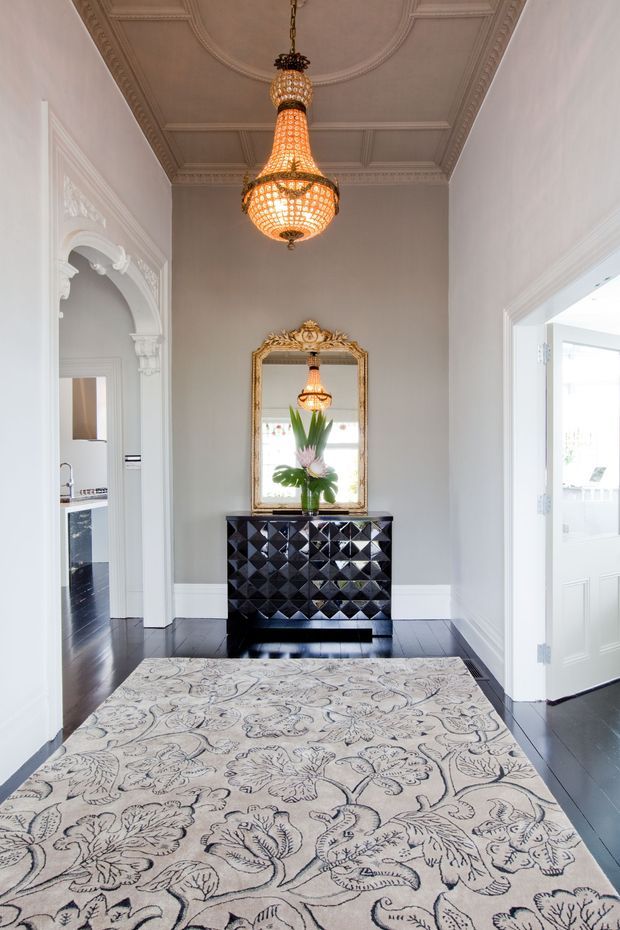 Elegant residential hallway or entry foyer with a pale grey wall, ornate white decorative mouldings, coffered ceiling, dark polished timber flooring, and a large patterned area rug. A black geometric-front console sits beneath an ornate gilded mirror, reflecting a decorative pendant light and floral arrangement. The space connects visibly to adjacent rooms through ornate archways and doorways.
