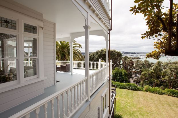 Exterior view of a white weatherboard home with a deep covered veranda, ornate white timber balustrades, decorative posts, multi-pane windows, and a landscaped garden overlooking a bay or coastal inlet. The image highlights the home's elevated veranda and waterfront outlook.