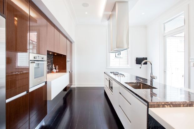Bright contemporary galley kitchen with dark glossy timber cabinetry along one wall, white integrated cabinetry and central island, dark timber flooring, and a dark polished stone benchtop. Stainless-steel appliances, undermount sink, pendant-style rangehood, white walls, and abundant natural light create a crisp high-contrast interior.