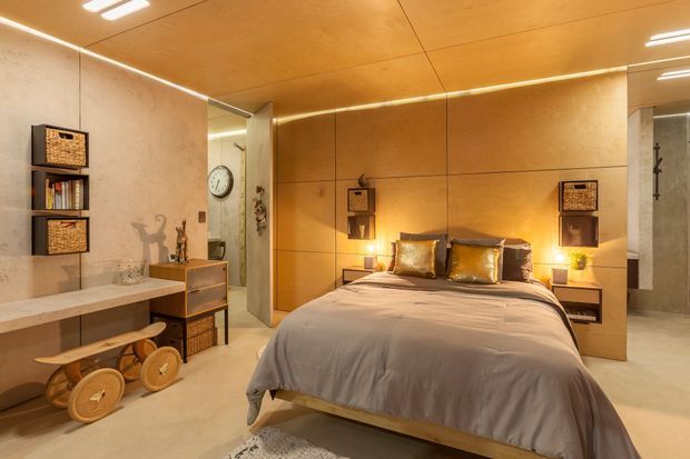 Warm, contemporary bedroom with a low platform bed, integrated timber feature wall and bedside niches, soft ambient lighting, built-in shelving, a desk or console, and a compact adjoining bathroom visible through the opening. The room uses natural wood tones, pale plaster or concrete-like surfaces, and understated furnishings.