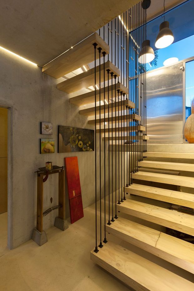 Interior view of a dramatic open-riser staircase with thick light timber treads suspended from the ceiling and supported by numerous slender black metal rods. The staircase rises through a tall space beside a textured concrete wall, with integrated warm lighting, pendant lights, artwork, and a bright glazed opening at the upper landing. The design has a sculptural, contemporary character.