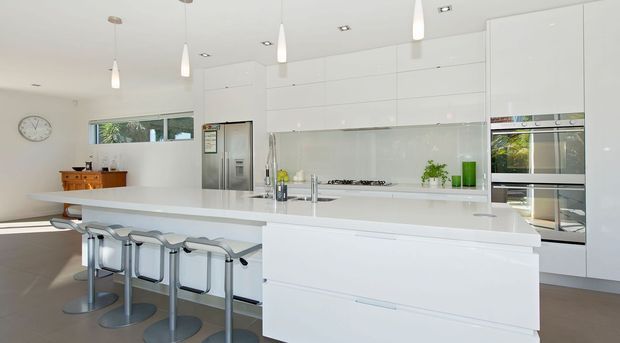 Bright contemporary white kitchen with a large central island and integrated breakfast-bar seating. The space features extensive glossy white cabinetry, a pale stone island benchtop, stainless-steel appliances, a light glass splashback, pendant lighting, and a small adjoining dining or living area.
