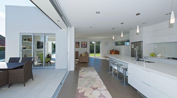 Bright open-plan contemporary kitchen and living area with a large white island, integrated white cabinetry, stainless-steel appliances, pendant lights, bar stools, and a dining area opening onto an outdoor terrace through expansive glazing. A lounge area and garden are visible beyond the kitchen.