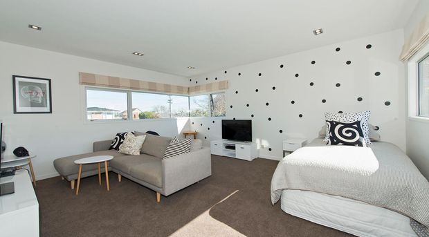 Bright contemporary open-plan bedroom and living area with a grey upholstered sofa, round timber coffee tables, a single bed, white media cabinetry, and a playful white feature wall covered in irregular black dots. Large windows provide natural light, while patterned cushions and artwork add graphic accents.