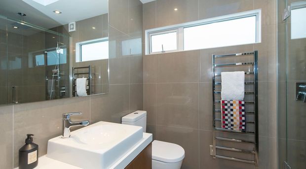 Bright contemporary bathroom featuring large taupe-grey wall tiles, a white vessel basin on a timber vanity, wall-mounted chrome tapware, a white toilet, frameless glass shower enclosure, mirrored cabinetry or wall mirror, and a chrome heated towel rail with folded towels. High horizontal windows provide natural light.