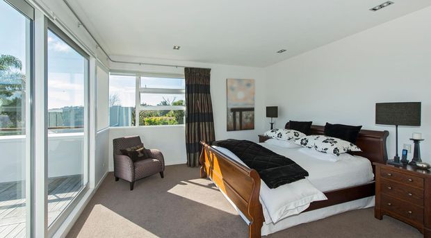 Bright contemporary bedroom with a large timber-framed bed, white bedding and black accents, matching wooden bedside furniture, neutral carpet, and extensive floor-to-ceiling glazing opening to a balcony or terrace. Natural daylight and a partial city or coastal outlook are visible through the windows.