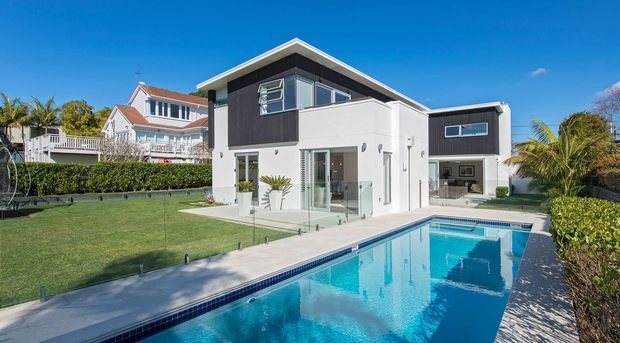 Exterior view of a contemporary two-storey residence with a crisp white and charcoal facade, low-pitched roof, large windows and glass balustrades. A paved terrace leads to a long rectangular swimming pool, with manicured lawn, hedges and palm planting around the property.