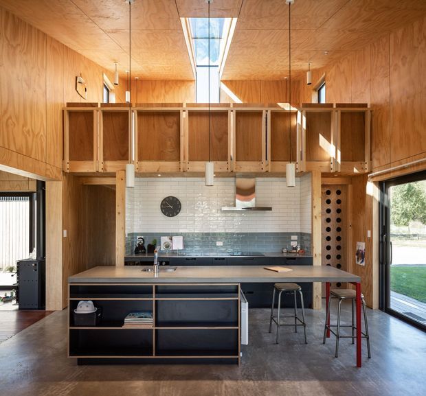 A warm, open-plan kitchen with extensive plywood wall and ceiling lining, a large island with dark open shelving, white subway-tile backsplash, dark blue-grey lower cabinetry, and a raised timber mezzanine or storage structure. A central skylight and clerestory glazing bring strong natural light into the double-height space, with bar stools at the island and a tall wine-storage niche beside the kitchen.