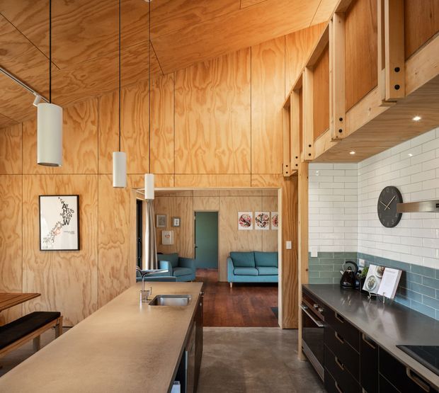 Warm open-plan kitchen viewed toward an adjoining living room, featuring extensive plywood wall and ceiling linings, a large island with an integrated sink, dark fixed cabinetry, a two-tone subway-tile splashback, timber structural framing and shelving, and suspended cylindrical pendant lights. The living area beyond contains blue sofas and a timber floor.