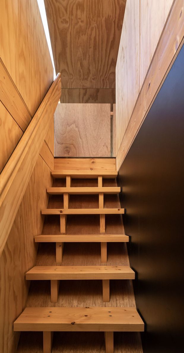 A narrow, steep interior stairway with open-riser timber treads and supporting vertical timber elements. Warm wood-panelled walls and a timber handrail line the left side, while a dark smooth wall creates contrast on the right. The stair rises toward a brighter upper landing and appears to use a compact, space-efficient design.