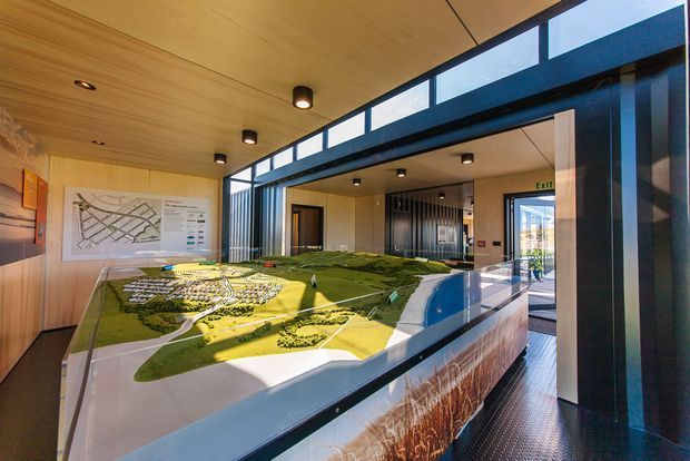 A contemporary architectural display space featuring a large illuminated landscape masterplan model on a central table. The room has extensive floor-to-ceiling glazing, warm timber ceiling and wall panelling, dark metal structural framing, recessed and surface-mounted lighting, and informational diagrams mounted on the wall. An entry or corridor is visible beyond the display.