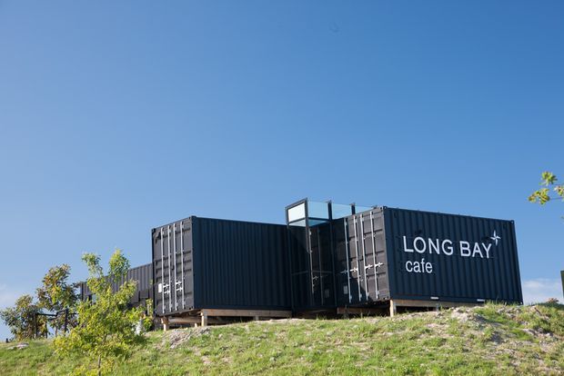 Exterior view of a contemporary café constructed from dark corrugated shipping containers, elevated on a sloping landscaped site. The building has large white signage reading “LONG BAY cafe,” dark metal container walls, glazed openings at the central junction, and a clear blue sky.