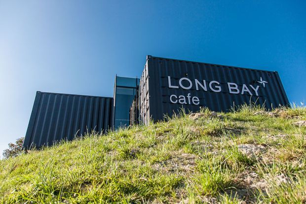 Exterior view of a cafe constructed from dark corrugated shipping-container-like modules, set into a grassy sloping site. The facade prominently displays the text “LONG BAY cafe,” with a bright blue sky behind it.