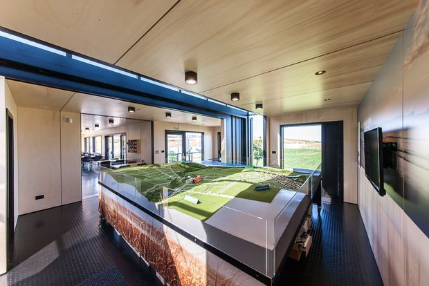 A bright contemporary exhibition or presentation room with a large illuminated architectural site model on a central display table. The space features pale timber wall and ceiling lining, dark ribbed flooring, recessed and cylindrical ceiling lights, extensive glazing, and a wall-mounted screen.