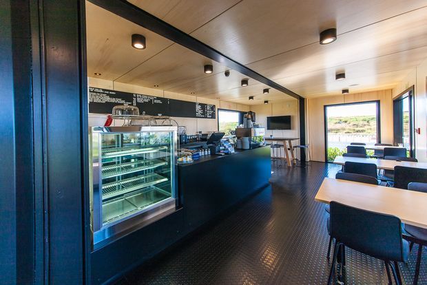 Interior view of a compact modern café or coffee shop, with a black service counter, refrigerated display case, espresso equipment, chalkboard menu, dining tables, and upholstered chairs. The space features warm timber ceiling panels, dark flooring and joinery, black-framed glazing, and contemporary pendant and recessed lighting.