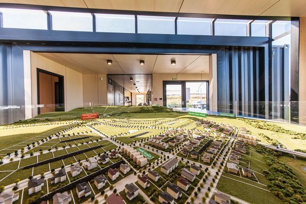 A large architectural scale model of a planned residential development is displayed on a table inside a bright, modern building. The model shows numerous houses, streets, landscaped areas, and open lots, with a glazed corridor and contemporary interior visible behind it.