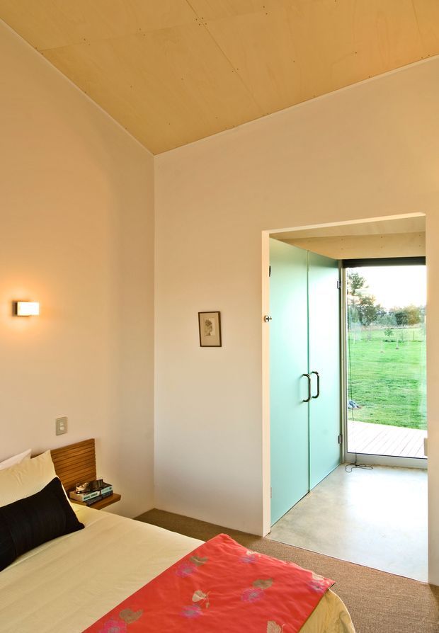 A compact bedroom with a partially visible bed, cream bedding, dark accent pillow and bright red patterned throw. The room has warm pale walls, a plywood-lined ceiling, a simple timber slatted headboard, wall-mounted light and a small framed artwork. A glass door opens directly to a deck and green garden, bringing in natural light.
