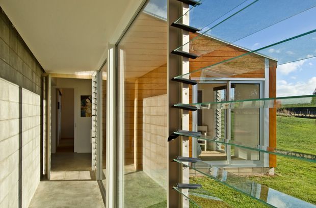 View from a narrow interior passage toward a glazed pavilion or extension. The space features exposed concrete block walls, polished concrete flooring, extensive floor-to-ceiling glazing, and distinctive horizontal glass louvres or fins in a dark metal frame. A warm timber-lined ceiling and a grassy exterior are visible beyond the glazing, with strong natural daylight throughout.