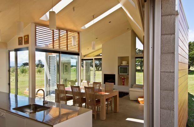 Bright open-plan kitchen and dining/living space viewed from a kitchen island. The room features a polished dark stone island benchtop with integrated sink, light cabinetry, a timber dining table and chairs, expansive glazing, timber horizontal louvres, a fireplace with built-in shelving, and a warm timber ceiling with multiple skylights or rooflights. The space opens onto a rural green landscape and has a relaxed contemporary architectural character.