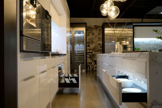 A high-end contemporary kitchen with glossy white full-height cabinetry, integrated appliances, a large white island with waterfall stone end, and a polished dark floor. A wine refrigerator, decorative textured feature wall, sculptural glass pendant lights, and illuminated open shelving add visual interest. The space has a dramatic, refined atmosphere with strong contrast between white joinery and dark ceiling and flooring.