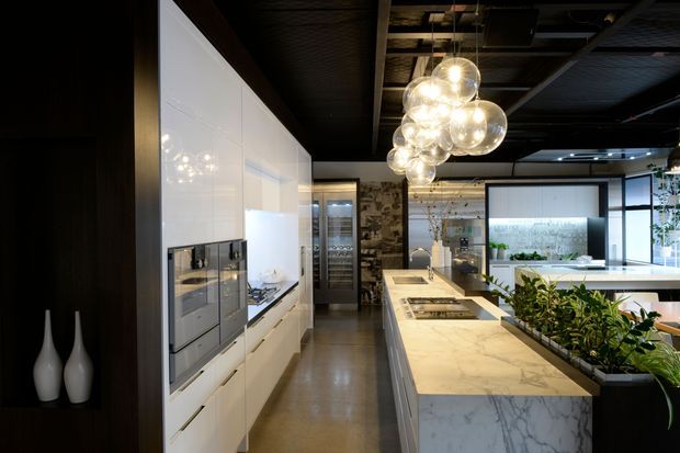 A sophisticated contemporary kitchen showroom or residential kitchen with a long white island topped in veined stone, integrated white cabinetry and appliances, a dark exposed ceiling, polished concrete floor, a planted trough, and a striking cluster of spherical pendant lights. A dining or living area is visible beyond the island.