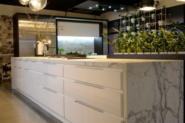 Contemporary kitchen showroom or large residential kitchen featuring a long white island with integrated drawers, a marble-look waterfall end panel and benchtop, dark ceiling cabinetry and display elements, pendant lighting, and a prominent built-in planter with lush greenery. A textured feature wall and illuminated rear display add visual interest.