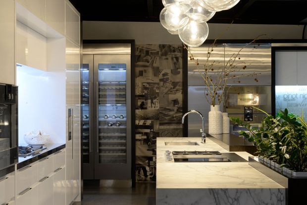A sophisticated contemporary kitchen with a large marble island, integrated white cabinetry, twin glass-front wine refrigerators, and a sculptural cluster pendant light. The space features a dramatic black ceiling, a monochrome patterned feature wall, a reflective splashback, and substantial indoor planting.